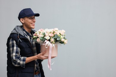 Photo of Smiling delivery man holding gift box with beautiful floral composition on grey background. Space for text