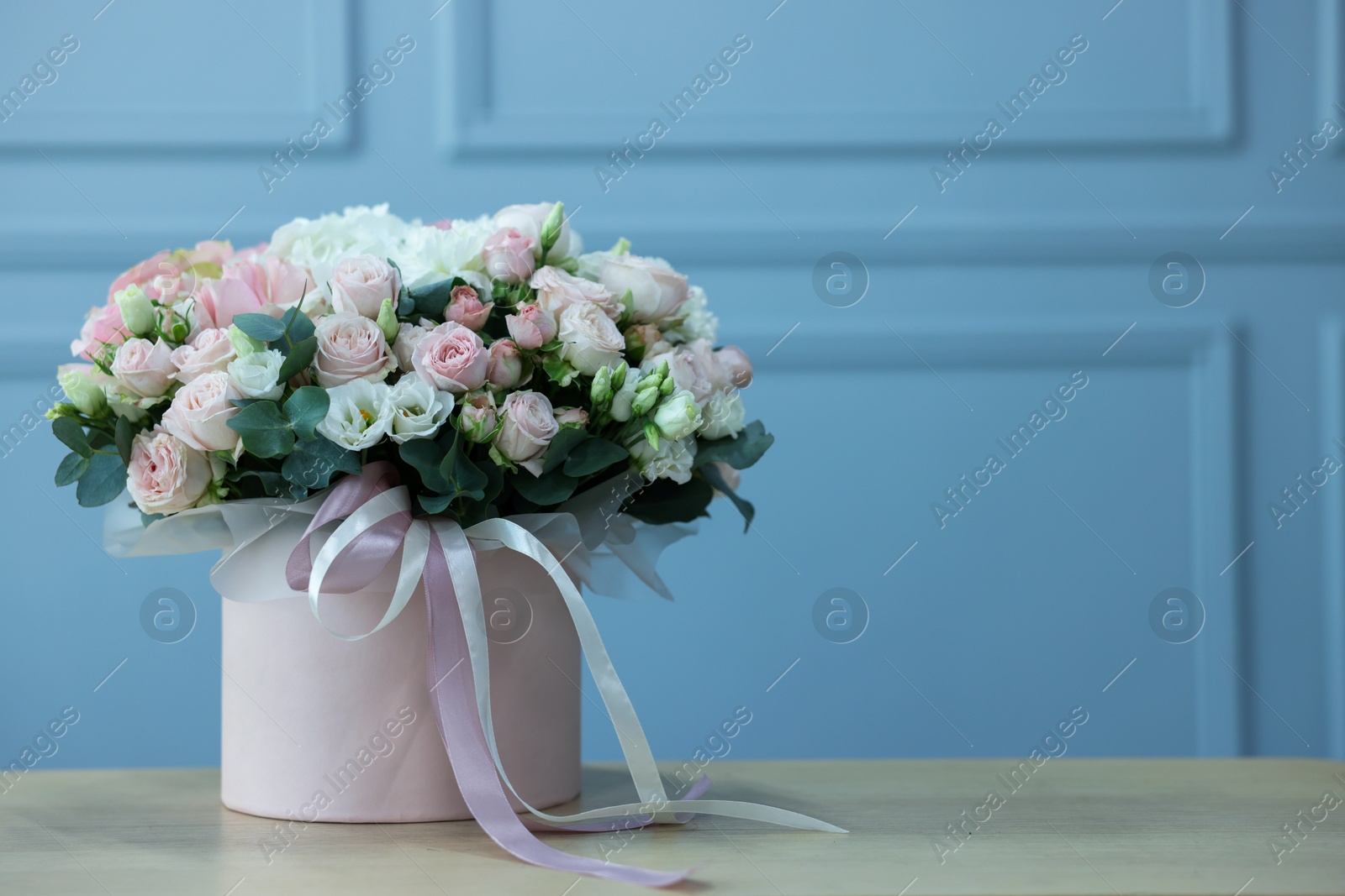 Photo of Gift box with bouquet of beautiful flowers and eucalyptus branches on wooden table against light blue wall. Space for text