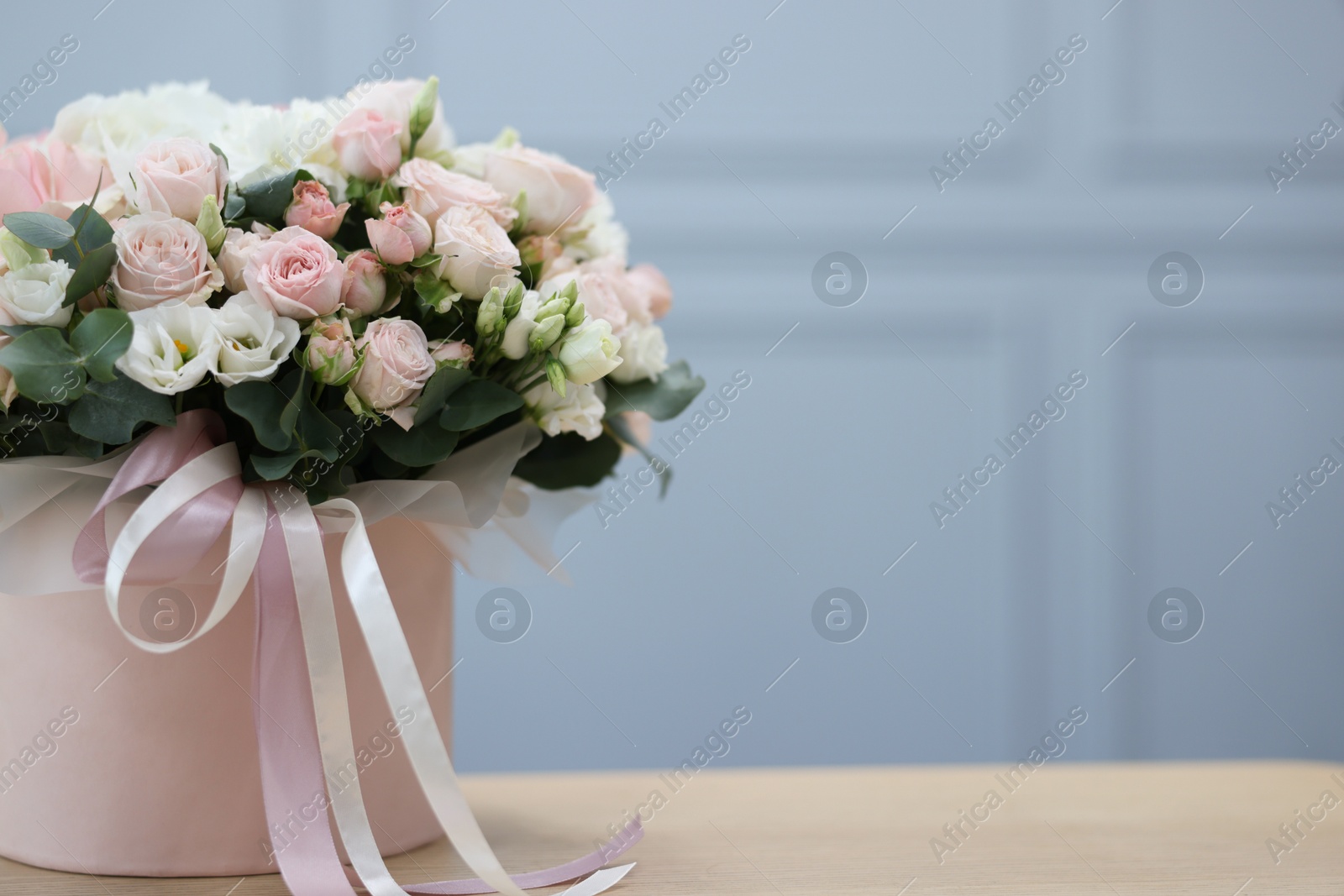 Photo of Gift box with bouquet of beautiful flowers and eucalyptus branches on wooden table against light blue wall, closeup. Space for text