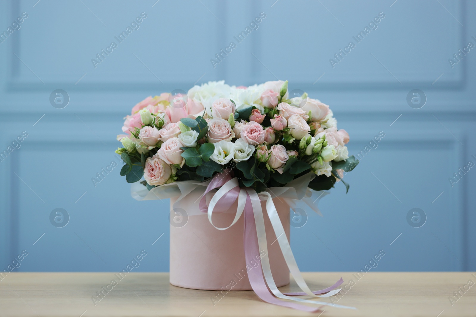 Photo of Gift box with bouquet of beautiful flowers and eucalyptus branches on wooden table against light blue wall