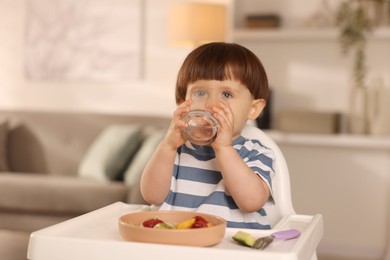 Photo of Cute little boy drinking water in high chair at home