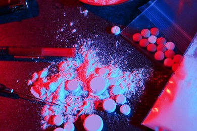 Photo of Drug addiction. Plastic bag with powder, pills and syringes on dark table in neon lights, flat lay