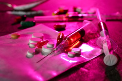 Photo of Drug addiction. Plastic bag with powder, syringes and pills on table in color light, closeup