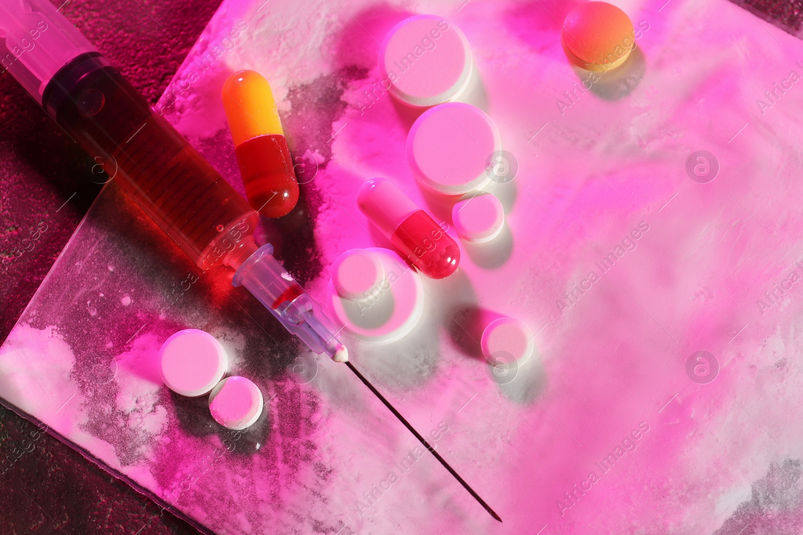 Photo of Drug addiction. Plastic bag with powder, syringe and pills on table in color light, top view