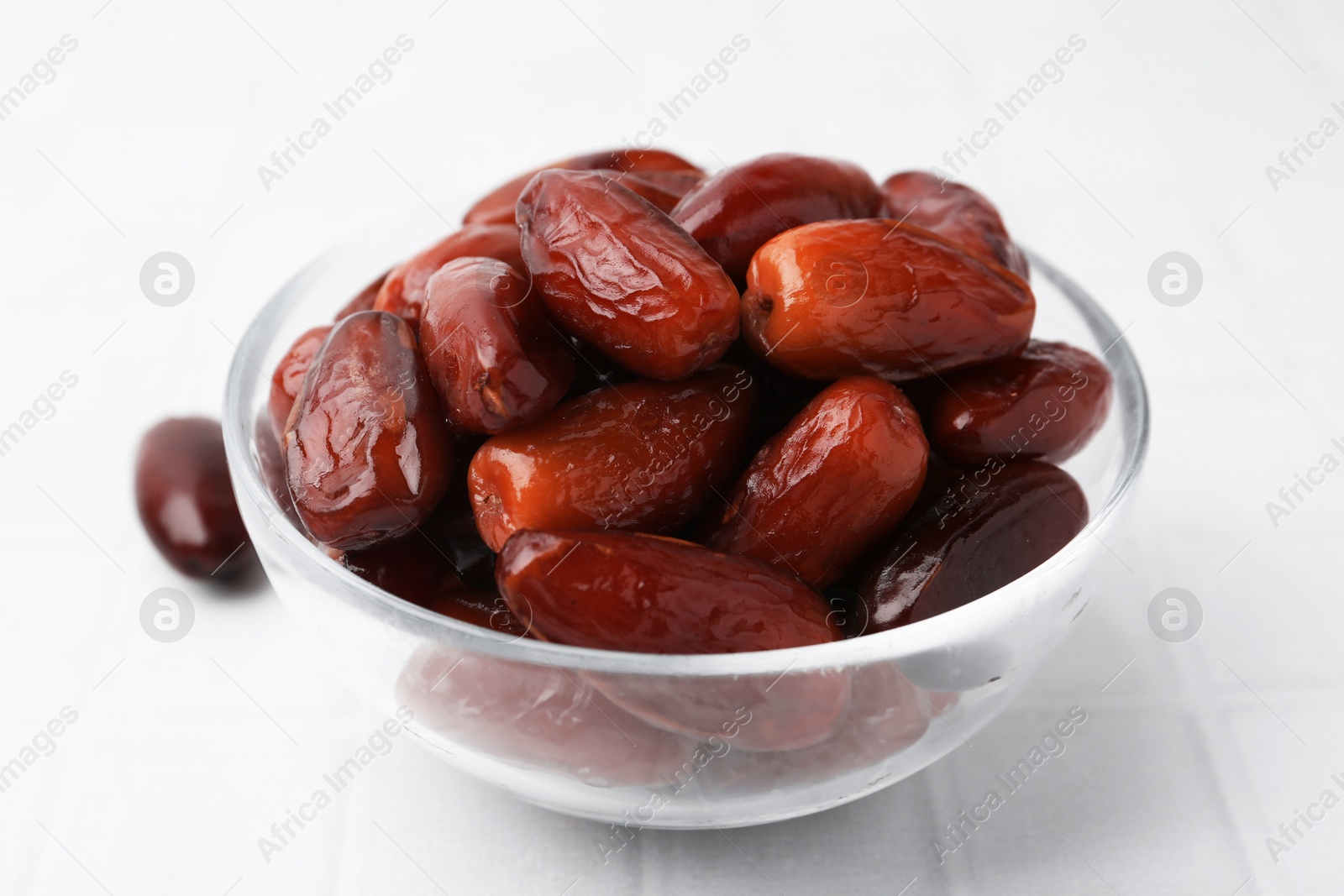 Photo of Tasty dried dates in glass bowl on white tiled table, closeup