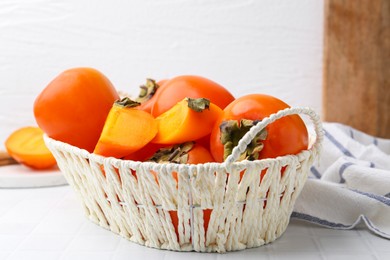 Photo of Ripe persimmons in wicker basket on white tiled table, closeup