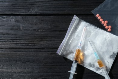 Photo of Drug addiction. Plastic bag with powder, pills and syringes on black wooden table, flat lay. Space for text