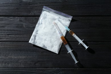 Photo of Drug addiction. Plastic bag with powder and syringes on black wooden table, top view