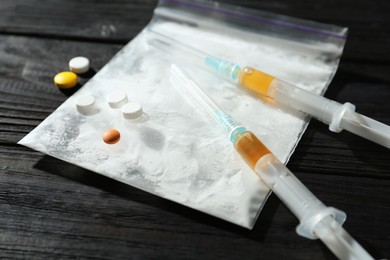 Photo of Drug addiction. Plastic bag with powder, syringes and pills on black wooden table, closeup