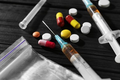 Photo of Drug addiction. Plastic bag with powder, syringes and pills on black wooden table, closeup