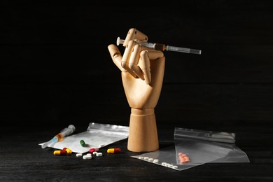 Photo of Drug addiction. Mannequin hand with syringe and pills on black wooden table against dark background