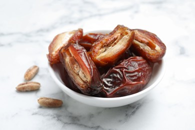 Photo of Many tasty dried dates with seeds in bowl on white marble table, closeup