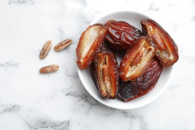 Photo of Many tasty dried dates with seeds in bowl on white marble table, top view