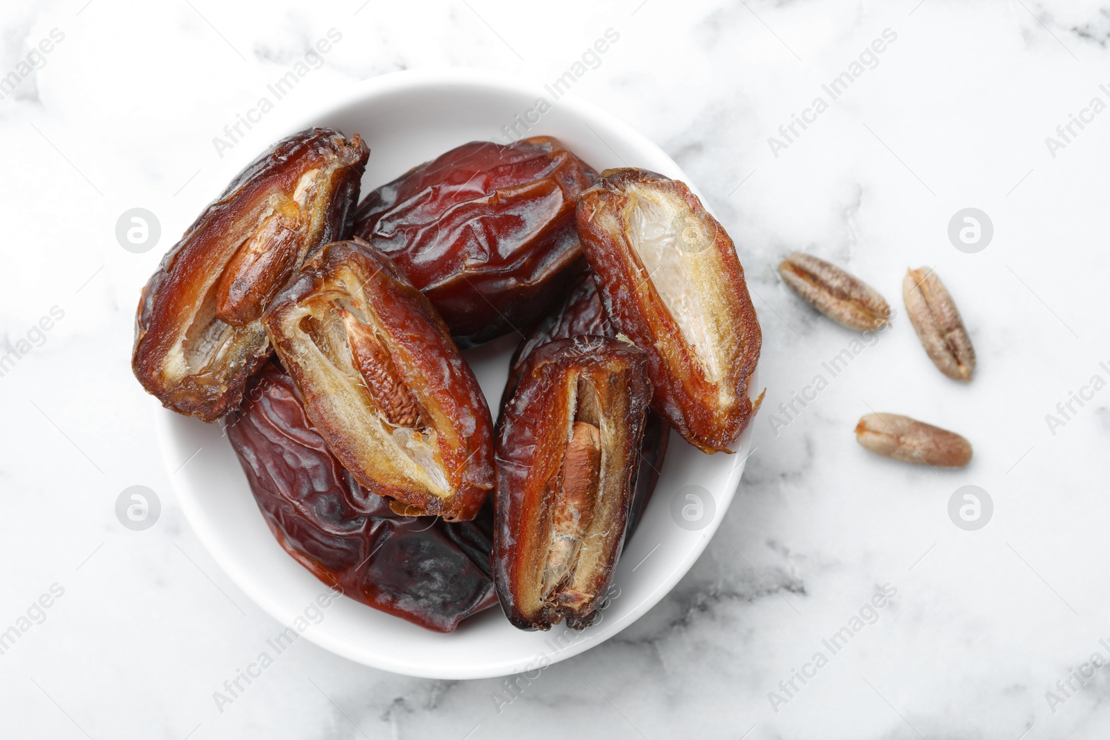 Photo of Many tasty dried dates with seeds in bowl on white marble table, top view