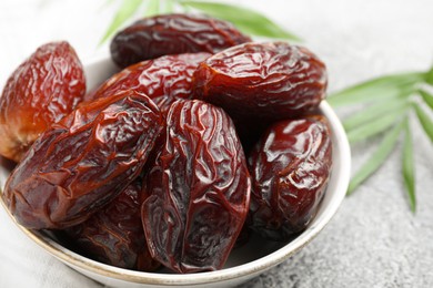 Photo of Many tasty dried dates in bowl and leaves on gray textured table, closeup