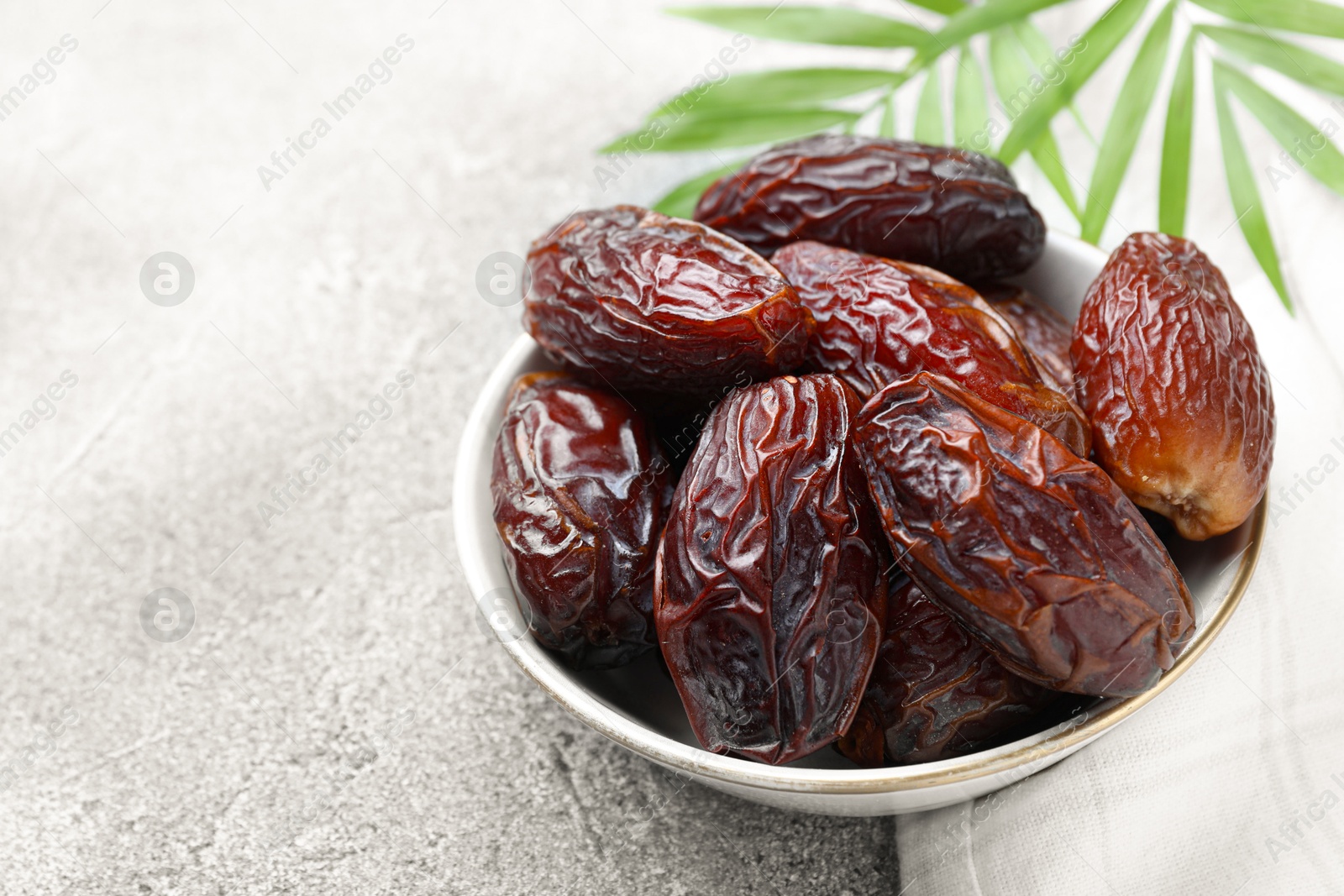 Photo of Many tasty dried dates in bowl and leaves on gray textured table, closeup. Space for text