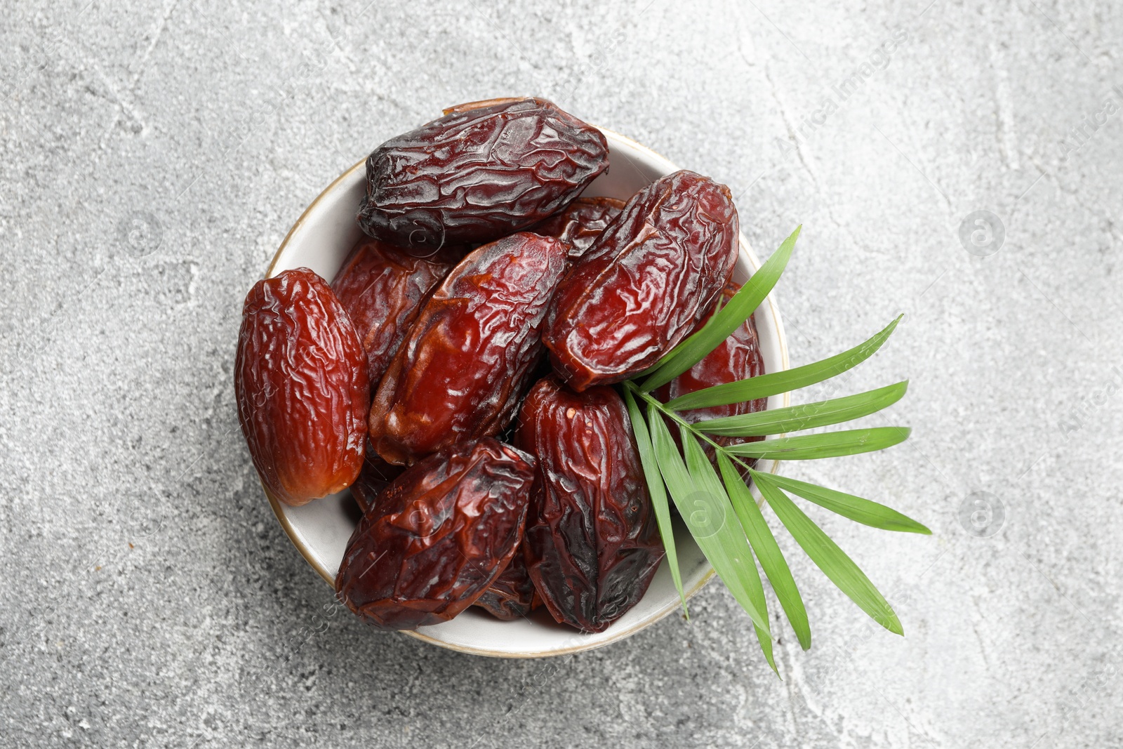 Photo of Many tasty dried dates and leaf in bowl on gray textured table, top view. Space for text