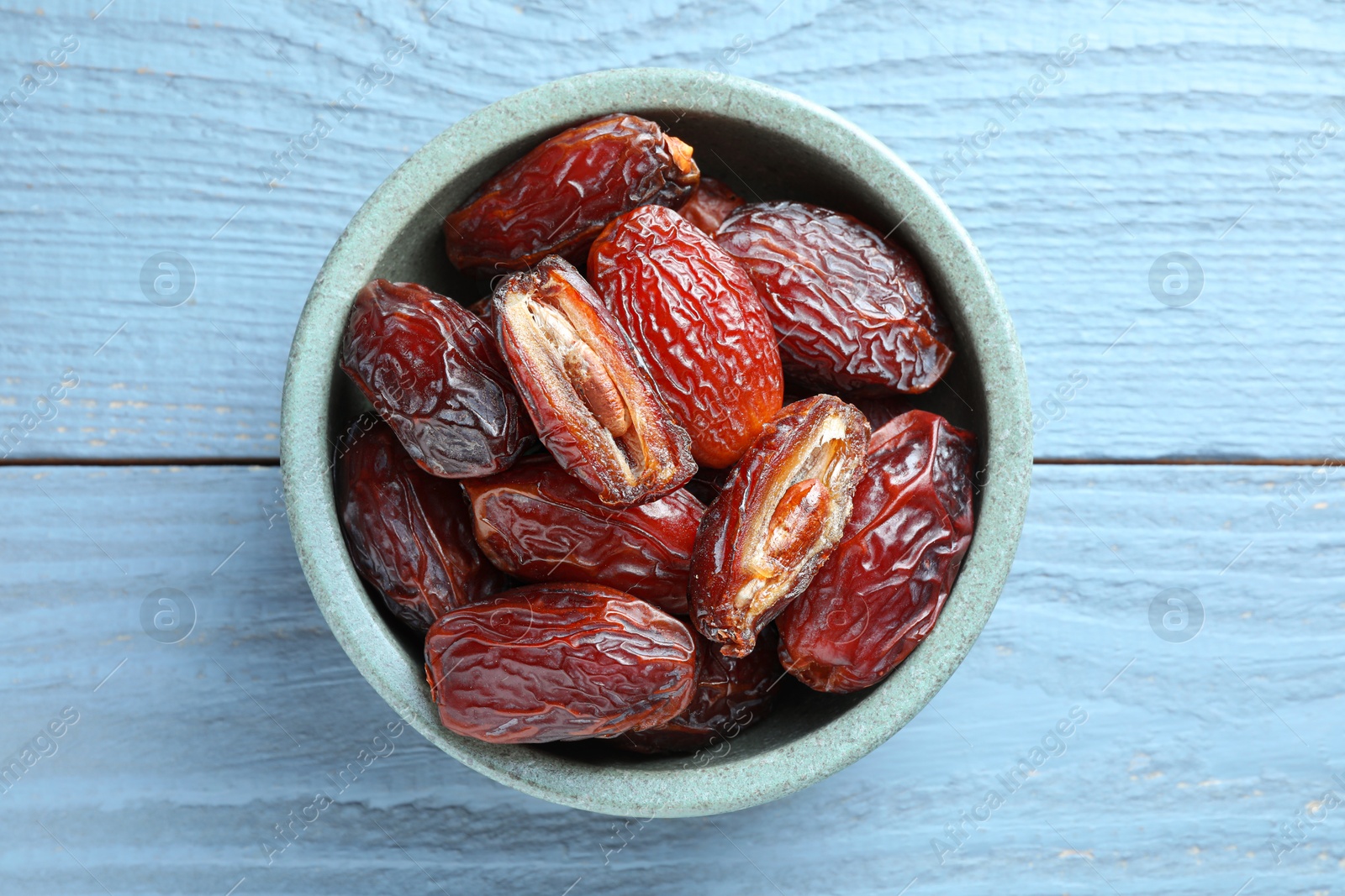 Photo of Many tasty dried dates in bowl on blue wooden table, top view