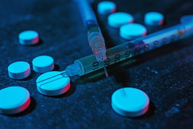 Photo of Drug addiction. Pills and syringes on dark textured table in neon lights, closeup