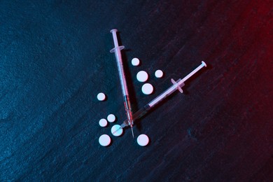 Photo of Drug addiction. Pills and syringes on dark textured table in neon lights, flat lay