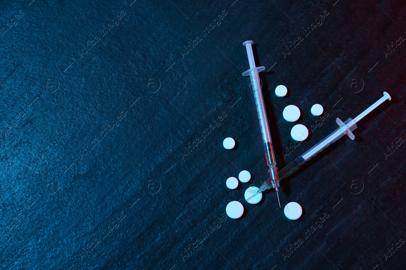 Photo of Drug addiction. Pills and syringes on dark textured table in neon lights, flat lay. Space for text