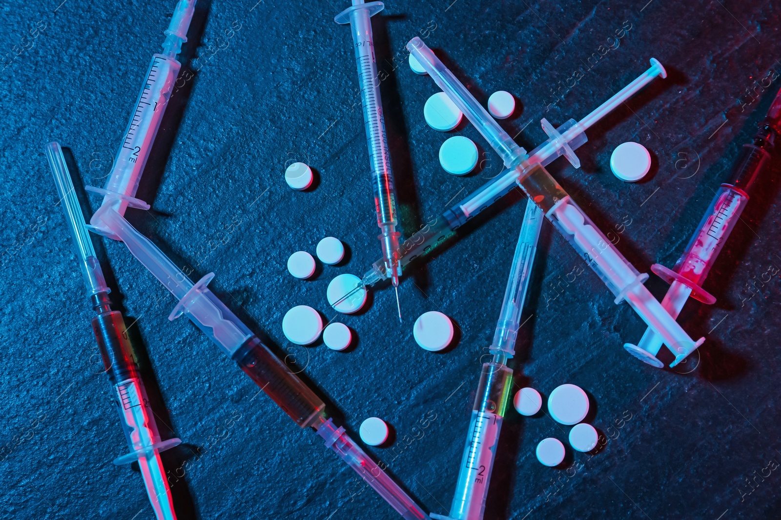 Photo of Drug addiction. Pills and syringes on dark textured table in neon lights, flat lay