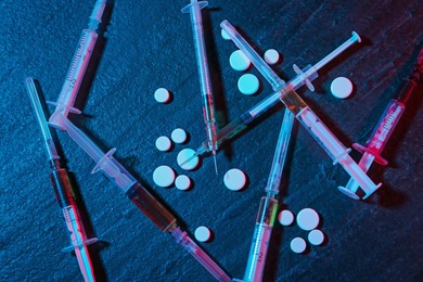 Photo of Drug addiction. Pills and syringes on dark textured table in neon lights, flat lay