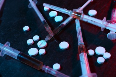 Photo of Drug addiction. Pills and syringes on dark textured table in neon lights, closeup