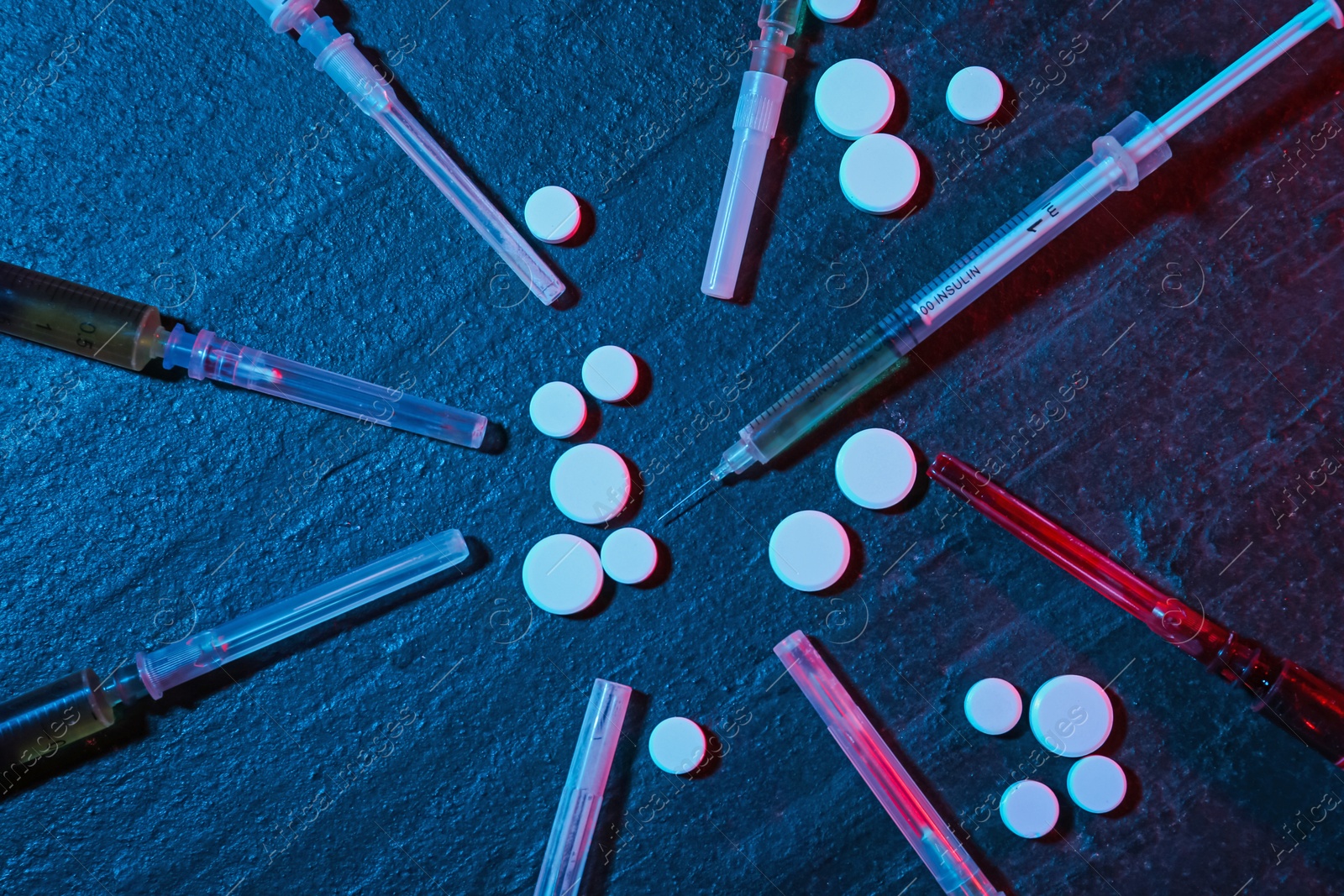 Photo of Drug addiction. Pills and syringes on dark textured table in neon lights, flat lay