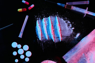 Photo of Drug addiction. Plastic bag with powder, pills and syringes on black table in neon lights, flat lay