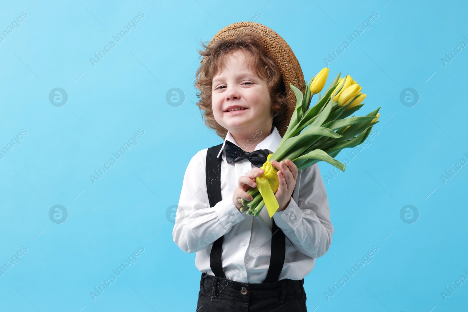 Photo of Cute little boy in wicker hat with bouquet of tulips on light blue background