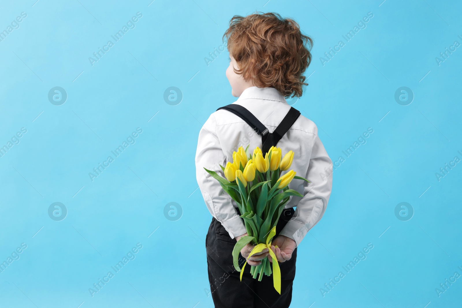 Photo of Little boy with bouquet of tulips behind his back on light blue background. Space for text