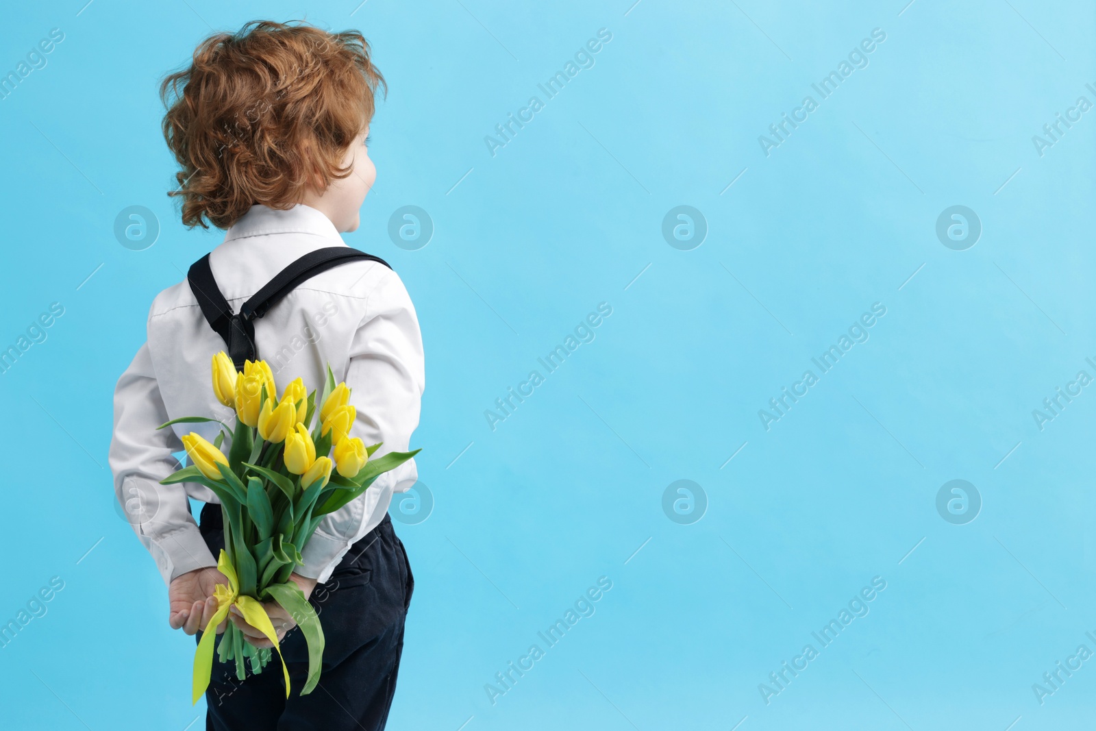 Photo of Little boy with bouquet of tulips behind his back on light blue background. Space for text