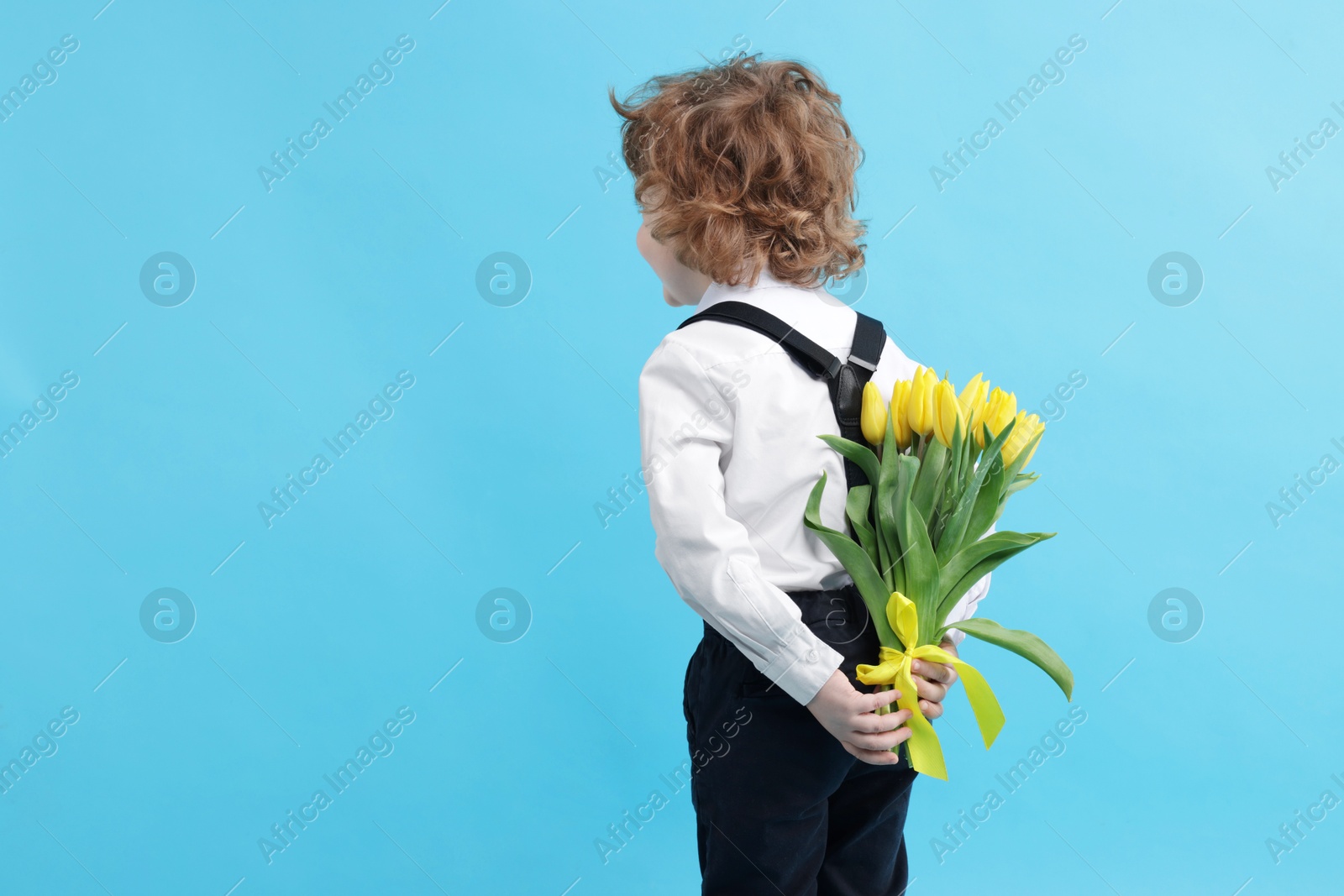 Photo of Little boy with bouquet of tulips behind his back on light blue background. Space for text