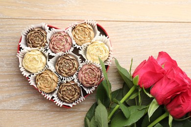 Photo of Flower shaped chocolate bonbons in box and red roses on wooden table, flat lay