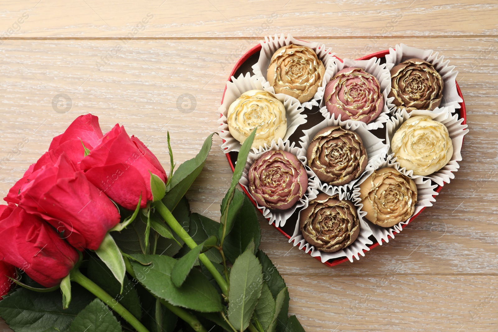 Photo of Flower shaped chocolate bonbons in box and red roses on wooden table, flat lay