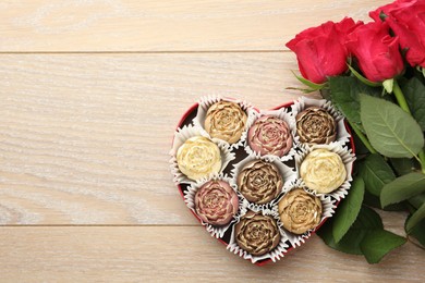 Photo of Flower shaped chocolate bonbons in box and red roses on wooden table, flat lay. Space for text