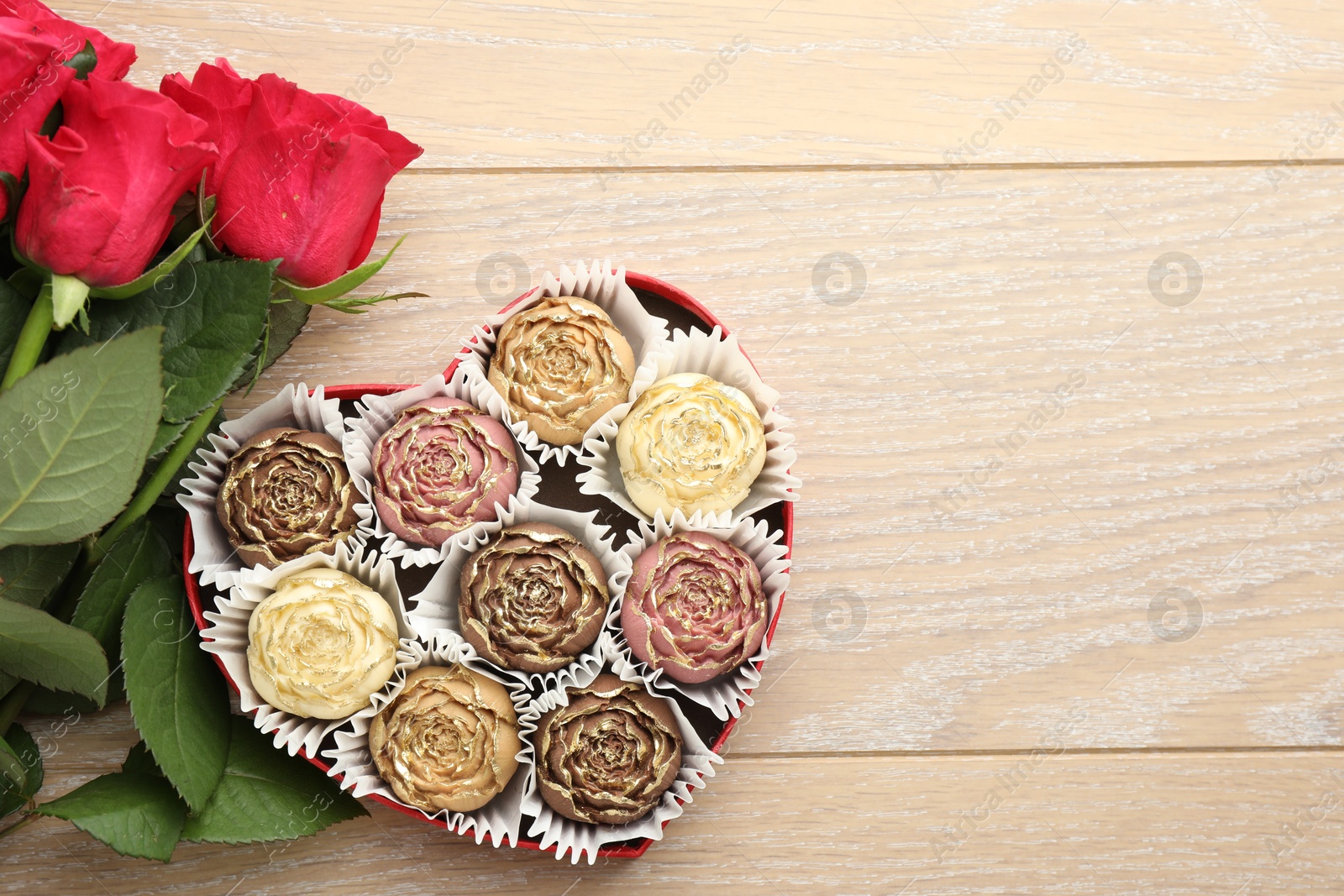 Photo of Flower shaped chocolate bonbons in box and red roses on wooden table, flat lay. Space for text