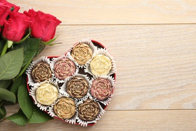 Photo of Flower shaped chocolate bonbons in box and red roses on wooden table, flat lay. Space for text