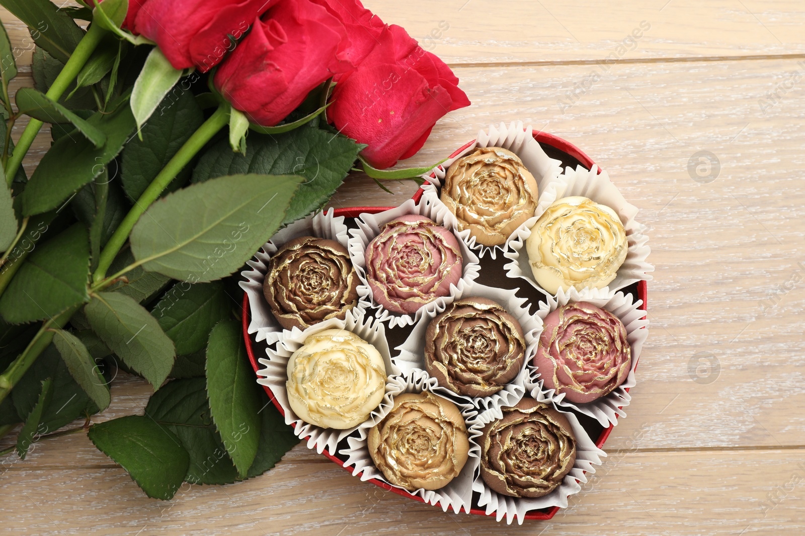 Photo of Flower shaped chocolate bonbons in box and red roses on wooden table, flat lay