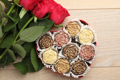 Photo of Flower shaped chocolate bonbons in box and red roses on wooden table, flat lay