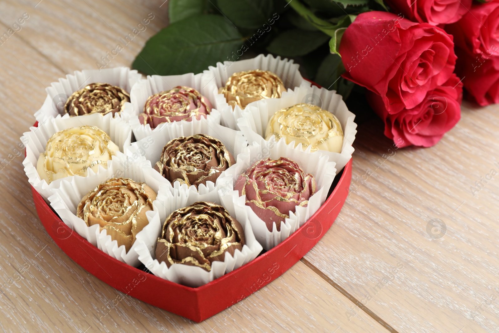 Photo of Flower shaped chocolate bonbons in box and red roses on wooden table, closeup