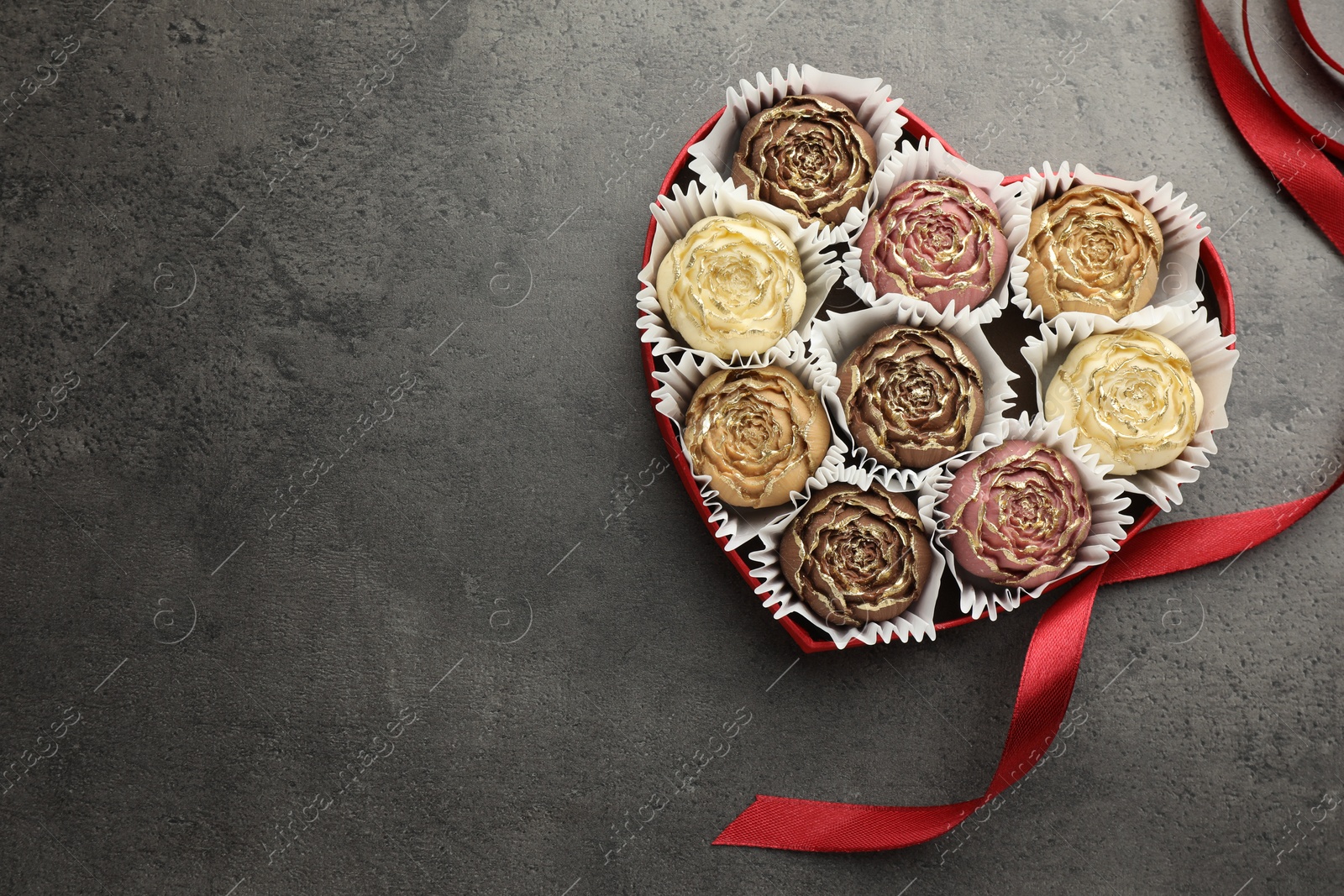 Photo of Flower shaped chocolate bonbons in box and red ribbon on grey table, top view. Space for text