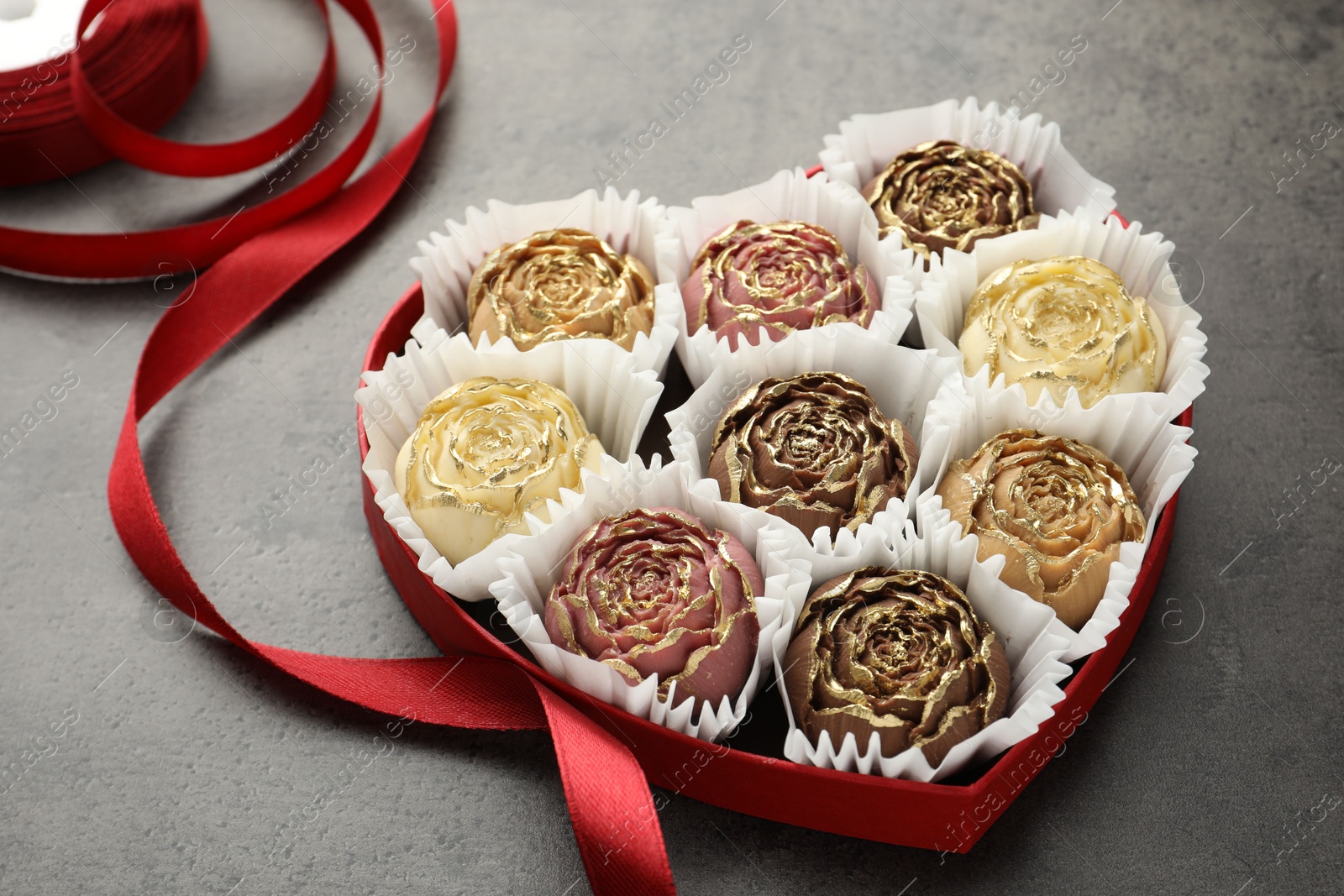 Photo of Flower shaped chocolate bonbons in box and red ribbon on grey table, closeup