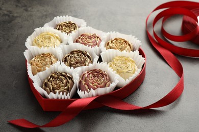 Photo of Flower shaped chocolate bonbons in box and red ribbon on grey table, closeup