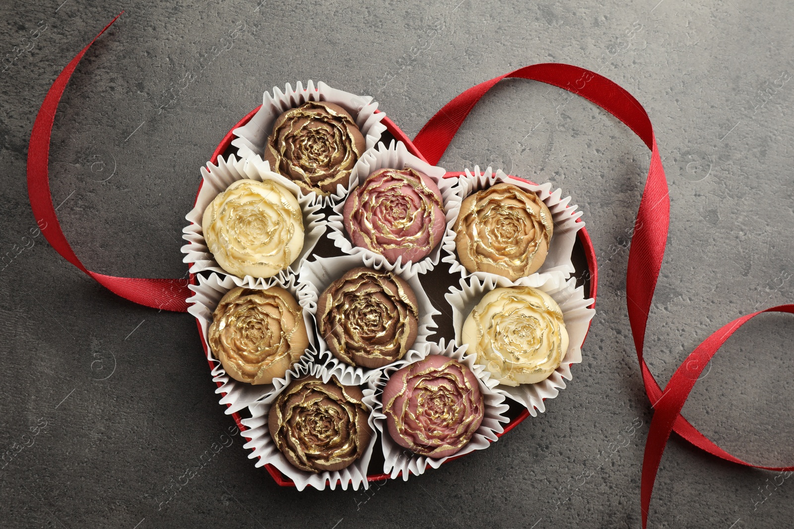 Photo of Flower shaped chocolate bonbons in box and red ribbon on grey table, top view