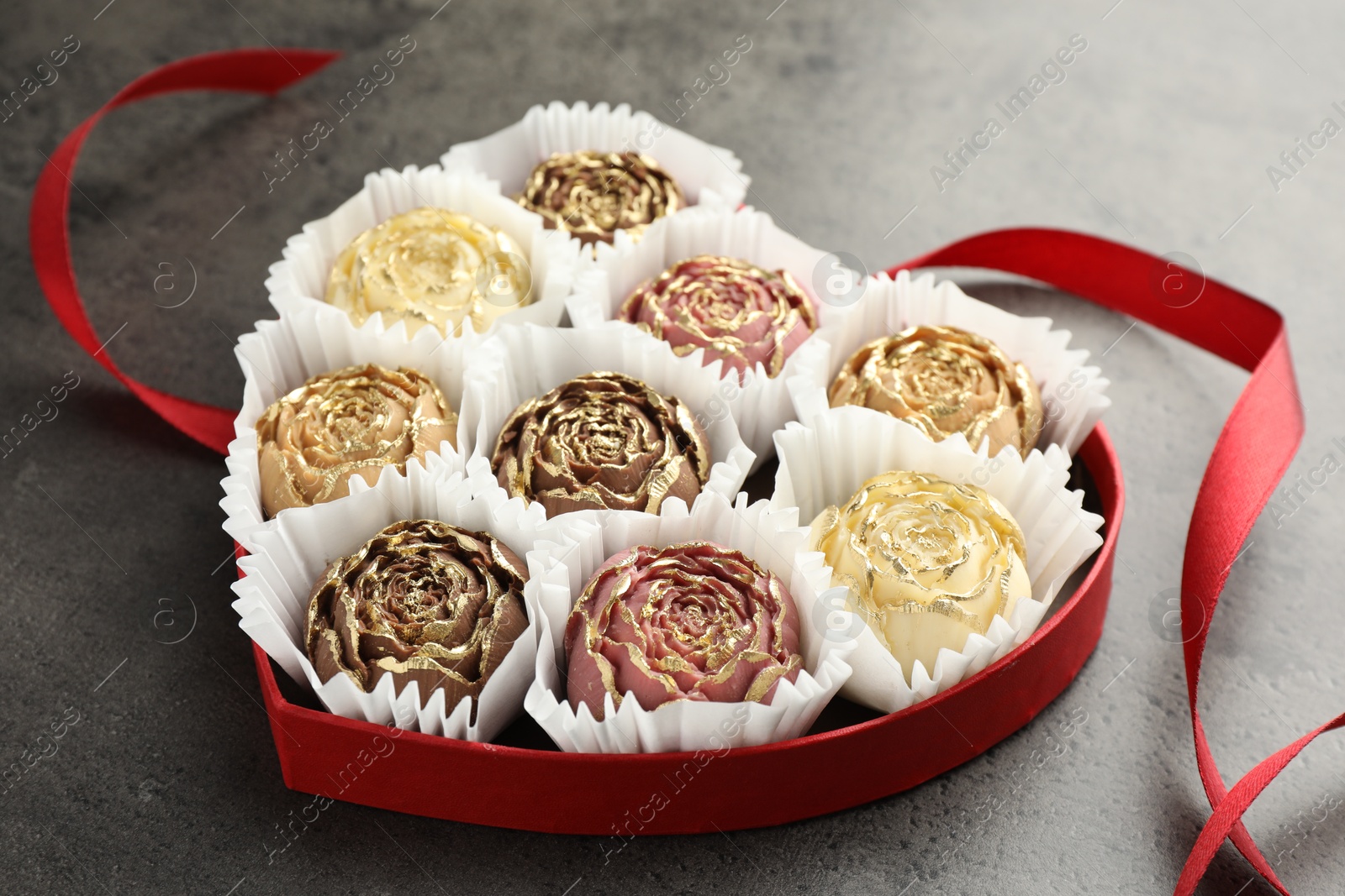 Photo of Flower shaped chocolate bonbons in box and red ribbon on grey table, closeup