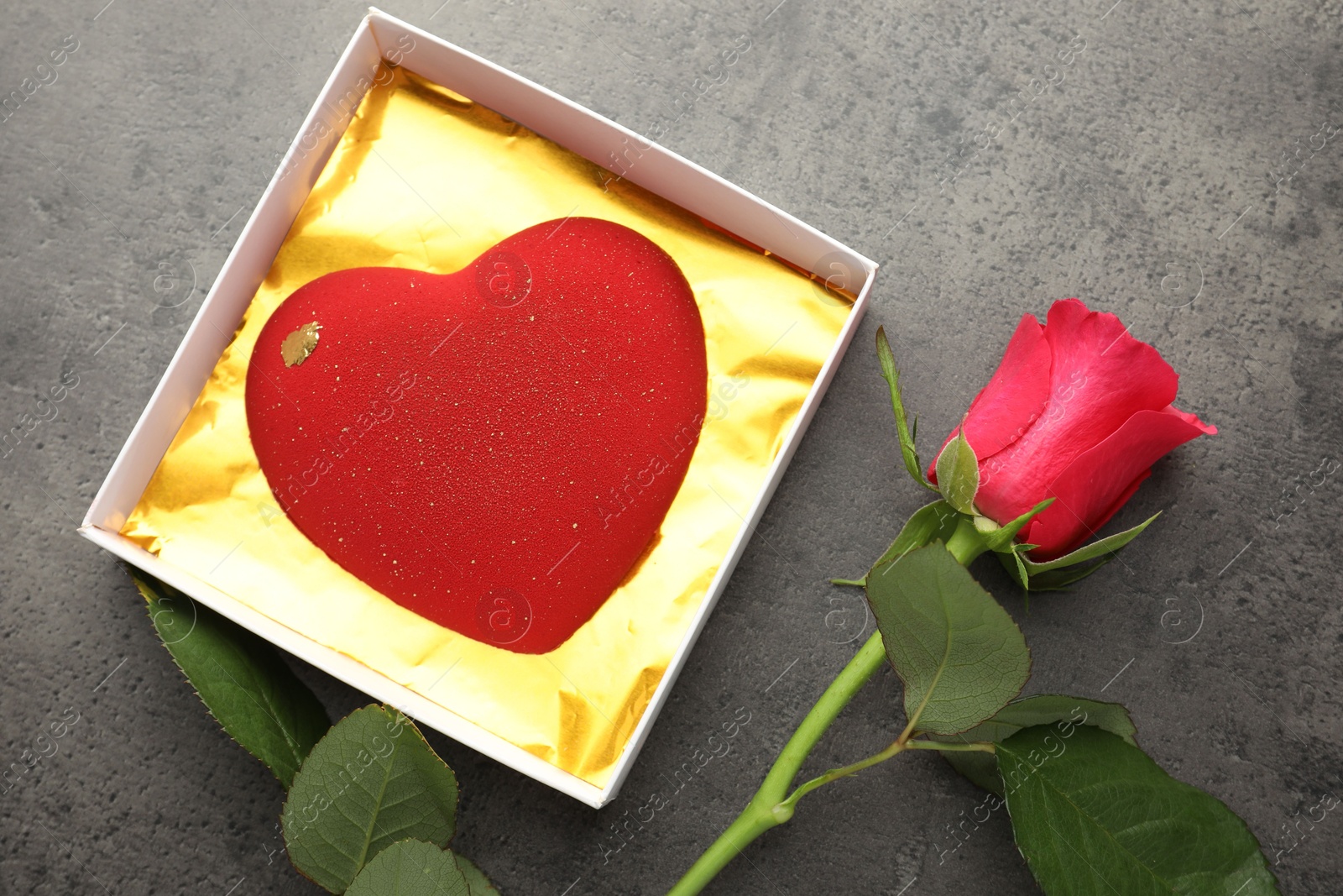 Photo of Heart shaped chocolate in box and red rose on grey table, flat lay