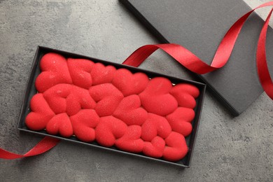 Photo of Chocolate bar decorated with hearts in box and red ribbon on grey table, flat lay