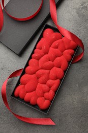 Photo of Chocolate bar decorated with hearts in box and red ribbon on grey table, flat lay